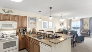 A modern kitchen with granite countertops, wooden cabinets, a white stove, and a dishwasher. There is a dining area with a table and chairs, and a living room with a couch and a view of the outdoors. - 14TH FLOOR OCEANVIEW CONDO|SHORT WALK TO THE BEACH UNIT 1408|ACCESS TO AMMENTIES (North Myrtle Beach)