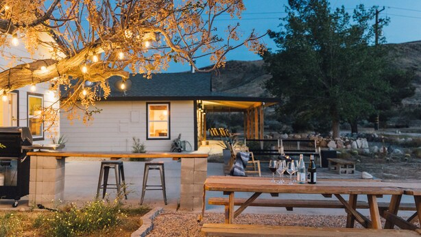 An outdoor dining area with a wooden table and benches, a barbecue grill, and string lights on a tree. - New Raven Ranch-Dreamy Compound With Spa, Hammock Circle, Yoga Platforms, & More (Yucca Valley)