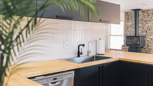 A modern kitchen with a wooden countertop, black cabinets, a built-in dishwasher, a sink, and a stone wall with a fireplace. - New Raven Ranch-Dreamy Compound With Spa, Hammock Circle, Yoga Platforms, & More (Yucca Valley)