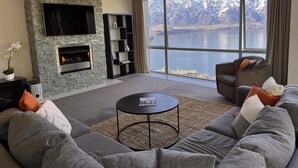 A modern living room with a stone fireplace, a large window with a mountain view, and a grey sectional sofa. - Iconic Queenstown Apartment (Queenstown)