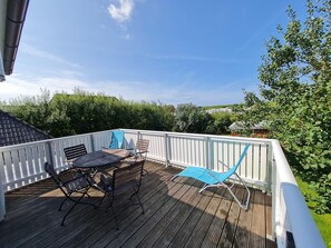 Um deck de madeira com guarda-corrimão branco, uma mesa, duas cadeiras e um toldo azul.