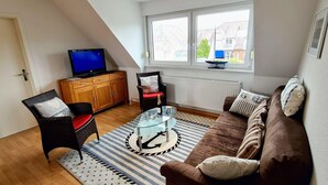 Un salon chaleureux avec un canapé marron, une table basse en verre ornée d’un motif d’ancre, un fauteuil en osier avec un coussin rouge et un téléviseur posé sur un meuble en bois.