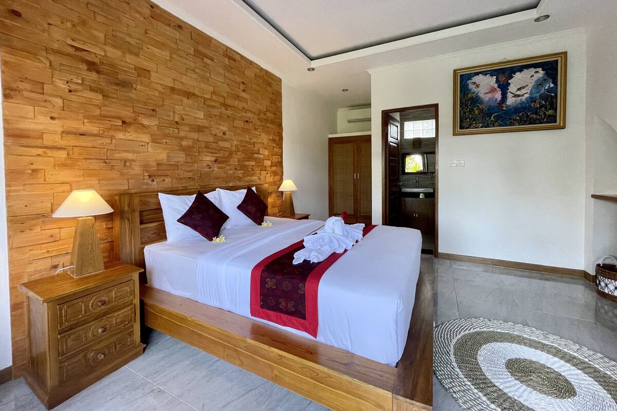A bedroom with a wooden headboard, a bed with white bedding and red accents, a nightstand with a lamp, a painting on the wall, and a view into the bathroom.