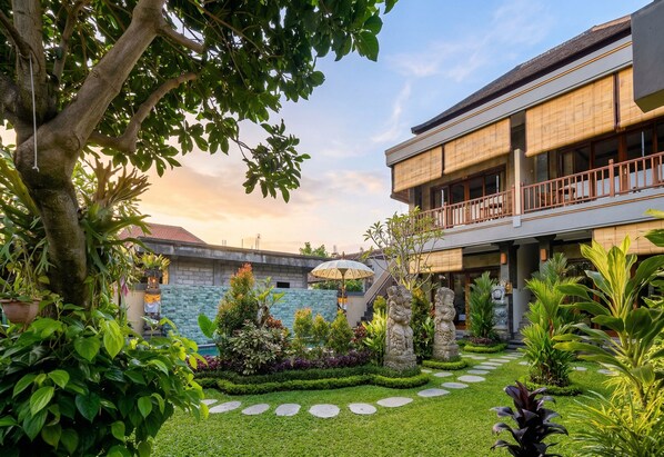 A courtyard with a stone pathway, lush greenery, and a building with a balcony. - Sari Murni (Ubud)