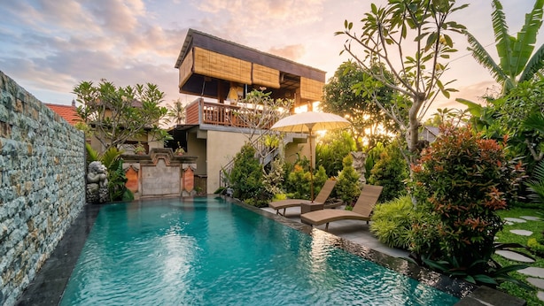 A swimming pool with lounge chairs, a stone wall, and a building with a balcony. - Sari Murni (Ubud)