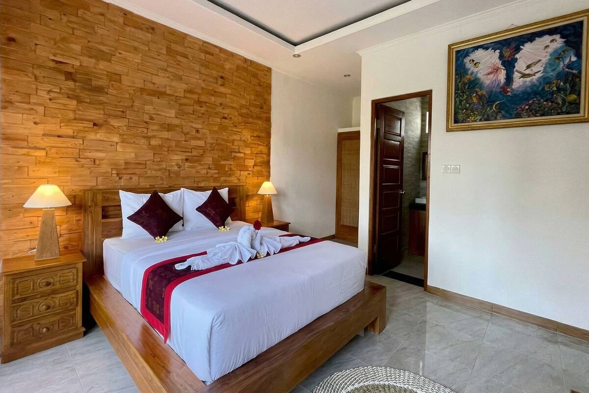 A hotel room with a wooden headboard, a bed with white linens and red accents, two bedside lamps, and a painting on the wall.