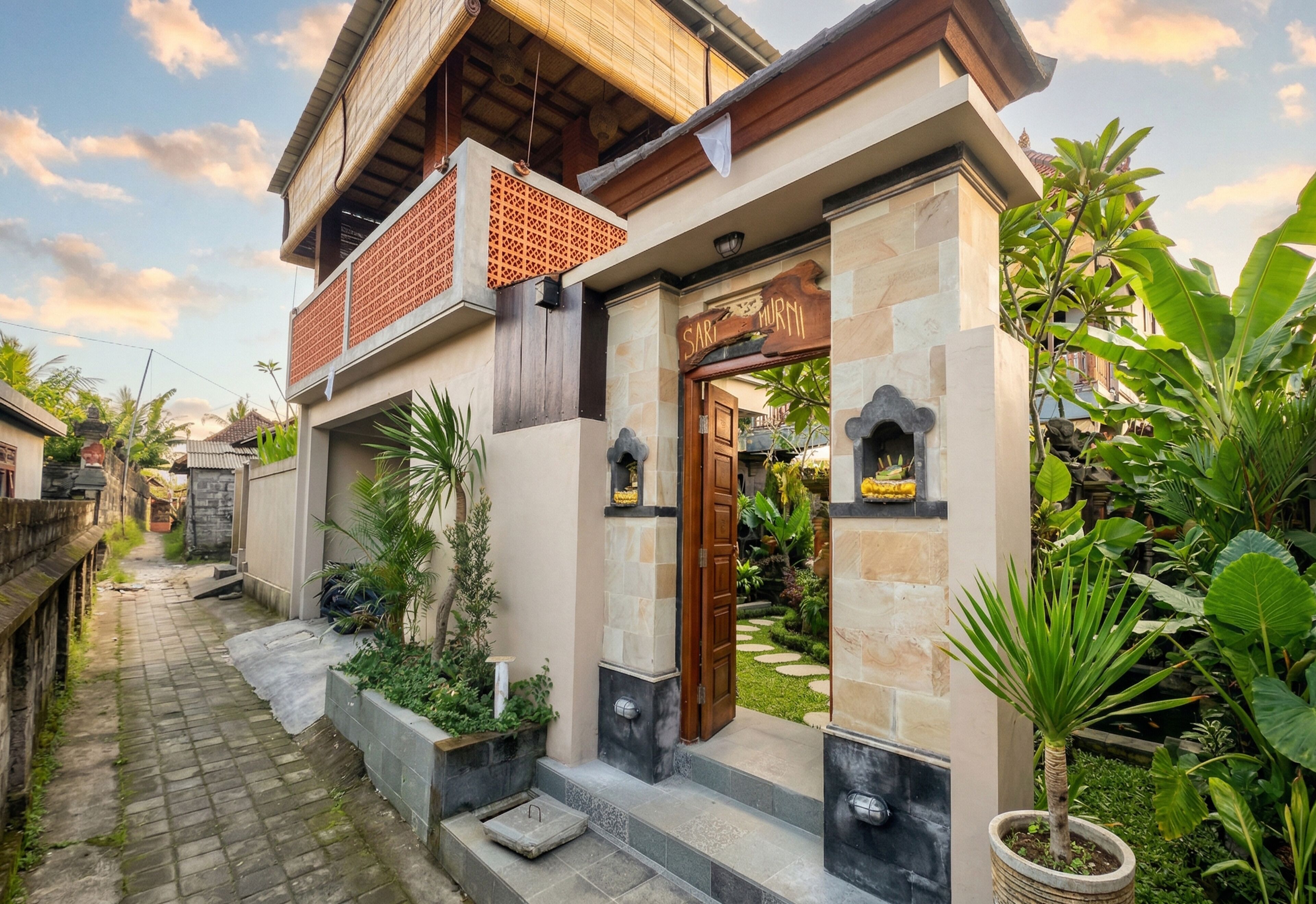 A two-story house with a stone facade, a wooden door, and a small garden with tropical plants.