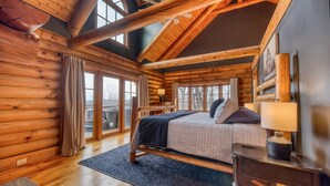 A log cabin bedroom with a bed, a nightstand, a lamp, and a view of the outside through large windows. - EPIC VIEWS-HotTub-WoodStove-Sauna-Fireplace-GameRoom-Playset-EV-FencedYard+More! (Rosman)