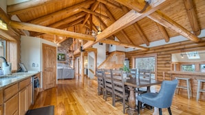 A spacious kitchen with wooden floors, a dining area with a table and chairs, and a high ceiling with exposed beams. - EPIC VIEWS-HotTub-WoodStove-Sauna-Fireplace-GameRoom-Playset-EV-FencedYard+More! (Rosman)