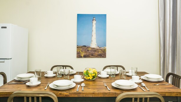 A dining table set with plates, cups, and a vase of flowers, with a white refrigerator and a painting of a lighthouse on the wall. - Aspid Studio & Apartment (Oranjestad)