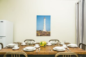 A dining table set with plates, cups, and a vase of flowers, with a white refrigerator and a painting of a lighthouse on the wall. - Aspid Studio & Apartment (Oranjestad)