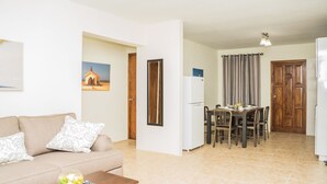 A living room with a sofa, coffee table, and a painting on the wall. There is a dining area with a table and chairs. - Aspid Studio & Apartment (Oranjestad)