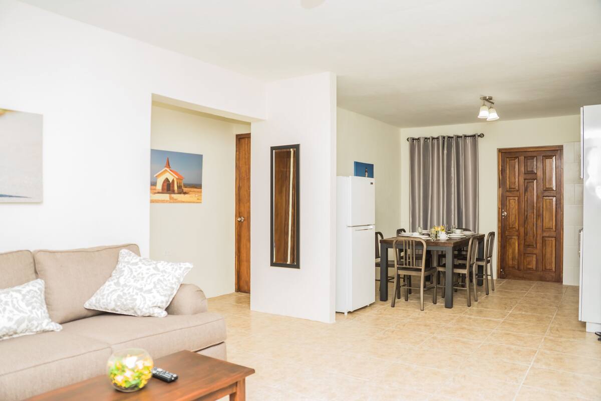 A living room with a sofa, coffee table, and a painting on the wall. There is a dining area with a table and chairs.