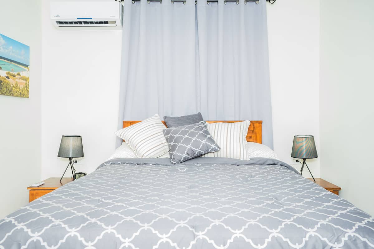 A bedroom with a bed, two bedside lamps, a wall-mounted air conditioner, and a curtain.