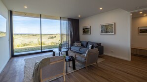 A spacious living room with a large window offering a view of a grassy landscape. There is a grey sofa, a wooden chair, and a small round table with a plant. - Links Lodge at The Dunes (Rye)