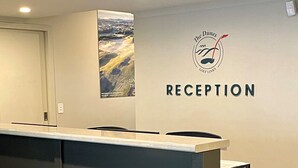 A reception desk with a logo above it, a wall-mounted poster, and a door to the left. - Links Lodge at The Dunes (Rye)