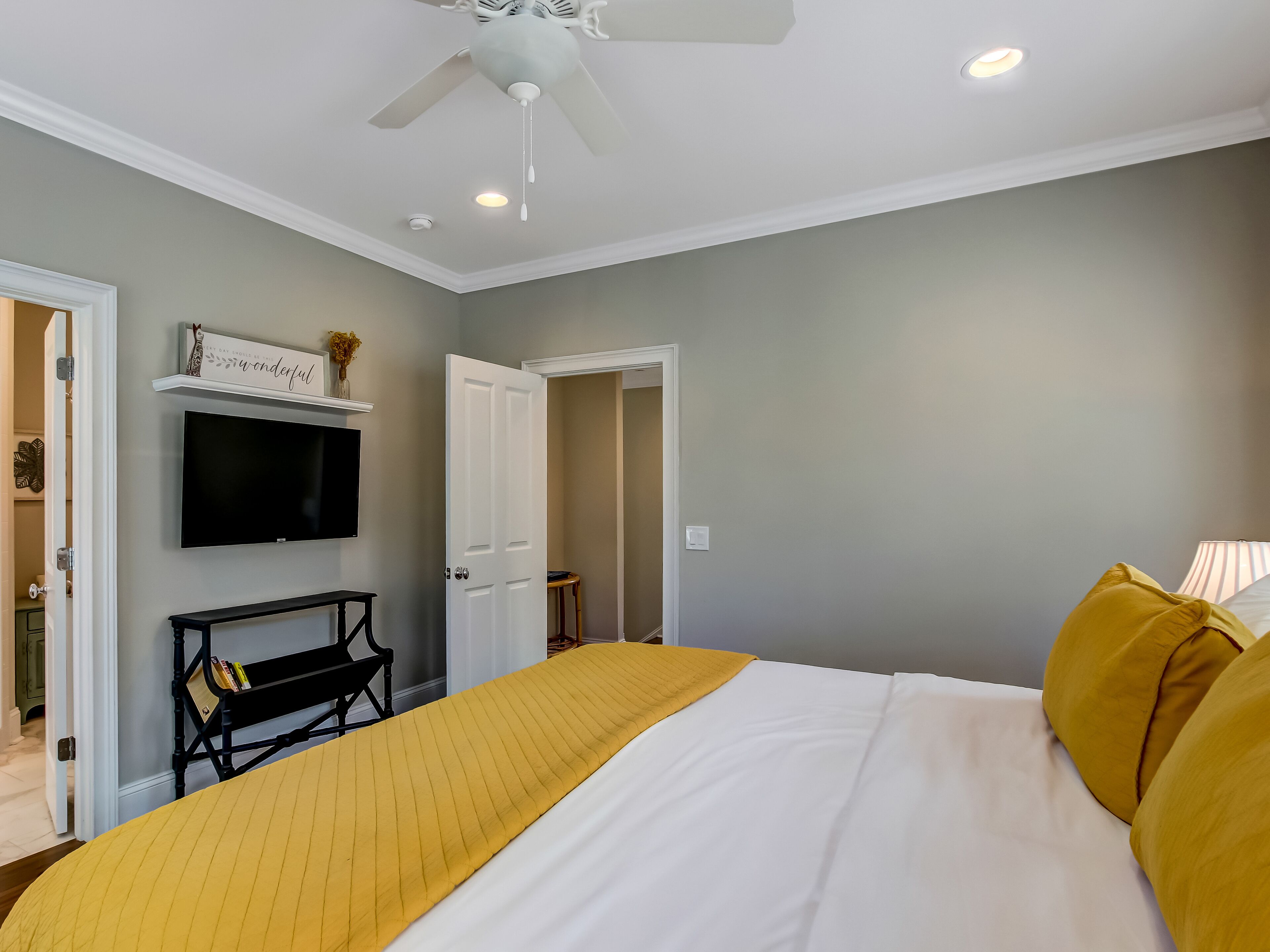 A bedroom with a bed, a ceiling fan, a TV mounted on the wall, and a door leading to another room.