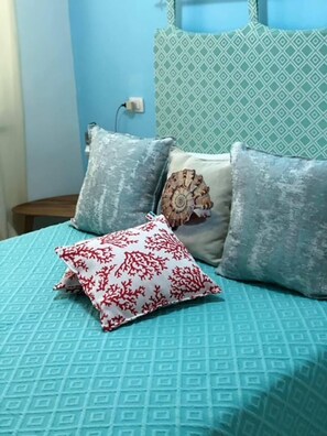 A bed with a blue patterned bedspread, three pillows, and a decorative shell. - Sweet Home ISOLA ROSSA (Trinita d'Agultu)