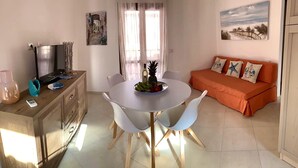 Ein Essbereich mit einem weißen Tisch, zwei weißen Stühlen, einer Obstschale und einer Weinflasche. Im Hintergrund ist ein Holzschrank mit einem Fernseher, einer Ananas und einer Fernbedienung zu sehen. Ein orangefarbenes Sofa mit Zierkissen bildet den Hintergrund.