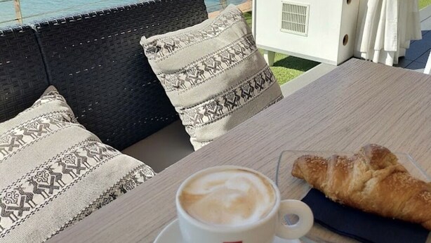 Ein Tisch mit einer Tasse Kaffee und einem Croissant, ein Korbsessel mit gemusterten Kissen und Blick auf das Meer und die Berge.