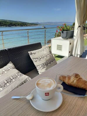 A table with a cup of coffee and a croissant, a wicker sofa with patterned cushions, and a view of the sea and mountains. - Sweet Home Mare LA TURRA (Trinita d'Agultu)
