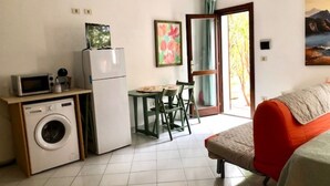 A compact living space with a washing machine, a small table with chairs, a bed, and a painting on the wall. - Sweet Home Mare Li FERULI (Trinita d'Agultu)
