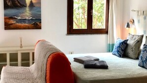 A bedroom with a bed, a window with curtains, a painting of a landscape, and a bedside lamp. - Sweet Home Mare Li FERULI (Trinita d'Agultu)