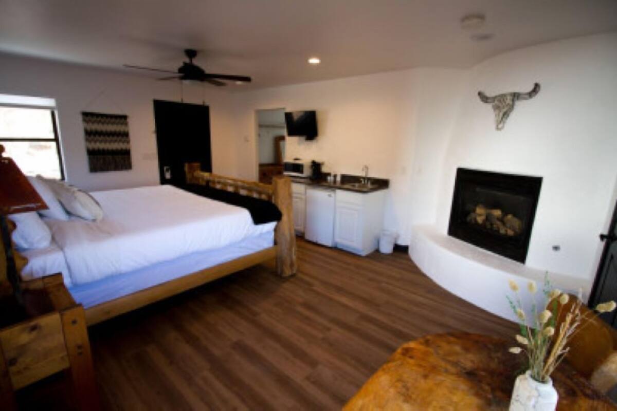 A bedroom with a wooden bed, a ceiling fan, a kitchen area, and a fireplace.