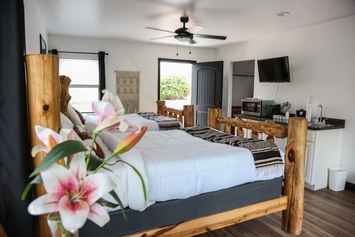 A bedroom with a wooden bed frame, a television, a desk with a computer, and a window with a view of greenery.