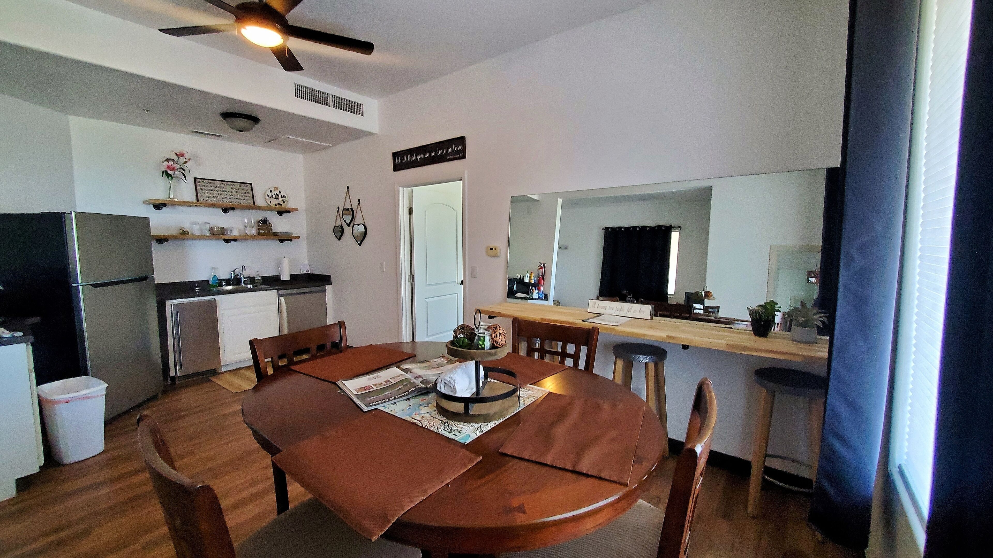 Eine kleine Küche mit Kühlschrank, Herd und Spüle. Ein Esstisch mit Stühlen und eine kleine Kücheninsel mit Schüssel und Krug sind vorhanden. Über dem Tisch ist ein Deckenventilator zu sehen.