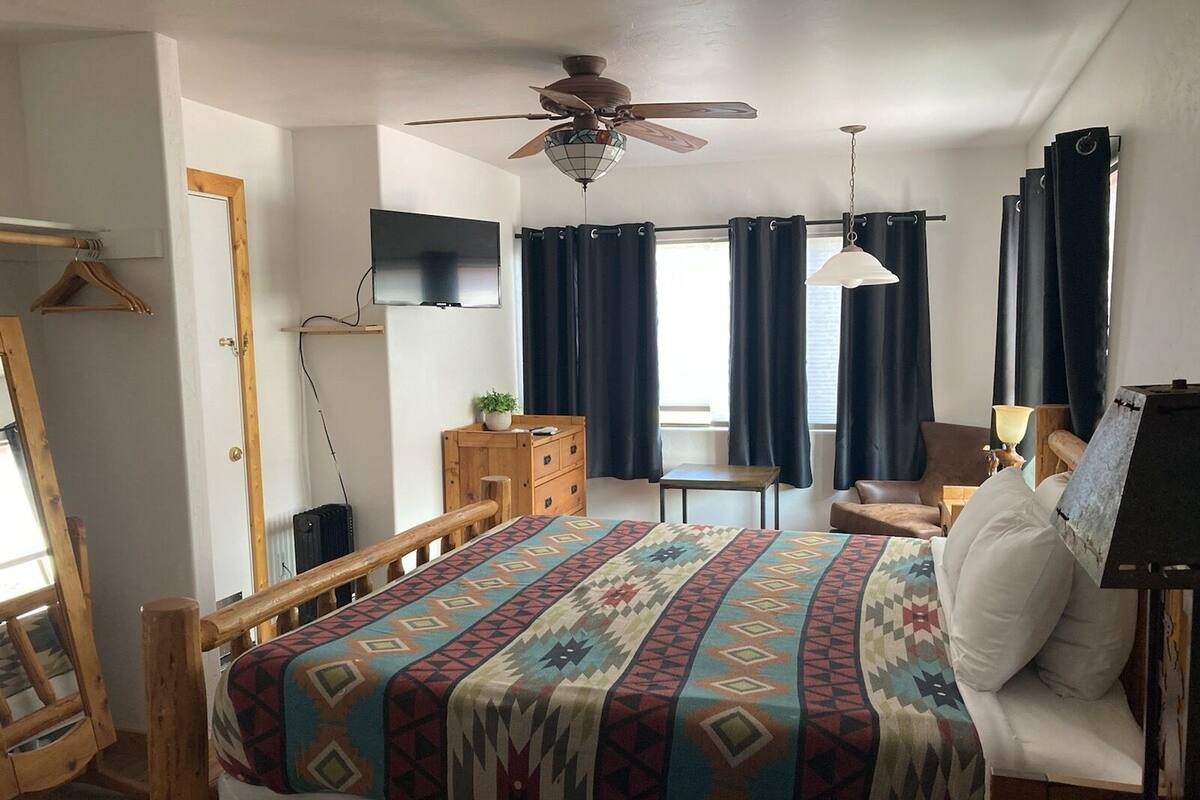 A bedroom with a bed, a wooden chair, a small table, a ceiling fan, and a TV mounted on the wall.