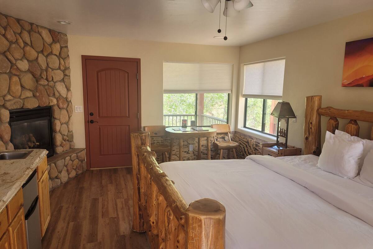 A bedroom with a stone fireplace, wooden headboard, and a view of greenery through large windows.