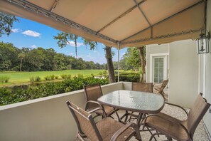 A covered outdoor patio with a glass table and wicker chairs, overlooking a green landscape and a body of water.
