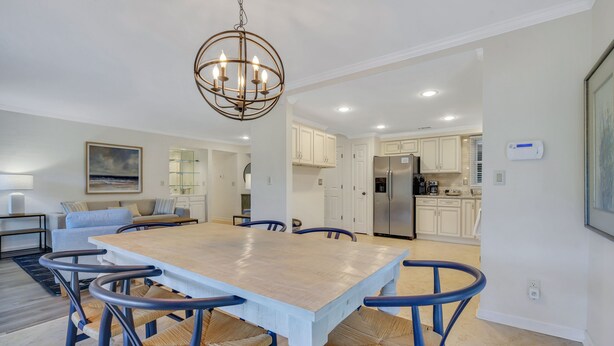 A spacious dining area with a large wooden table and blue chairs, a chandelier, and a view into the kitchen. - 276 Evian: Newly renovated single story unit with golf course views and tennis court access! (Hilton Head Island)