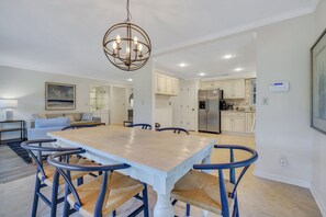 A spacious dining area with a large wooden table and blue chairs, a chandelier, and a view into the kitchen. - 276 Evian: Newly renovated single story unit with golf course views and tennis court access! (Hilton Head Island)