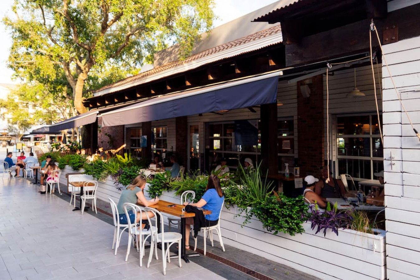 Un café en plein air, avec des tables et des chaises, entouré de verdure et bordé d’un bâtiment dont le toit est recouvert de tuiles.
