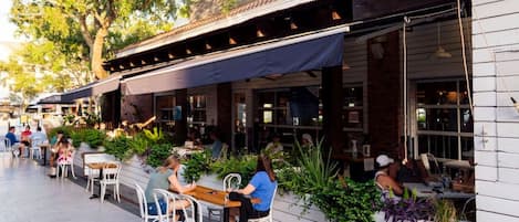 Un café al aire libre con mesas y sillas, rodeado de vegetación y un edificio con techo de tejas.