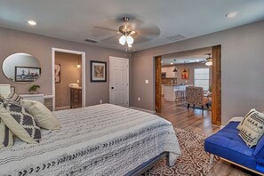 Un dormitorio con cama, mesitas de noche, un espejo y una puerta que da a la cocina y a la sala de estar.