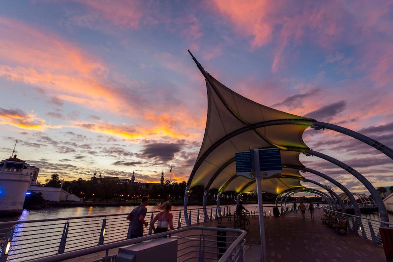 A waterfront promenade with a distinctive modern canopy structure, a boat docked on the left, and a calm river.
