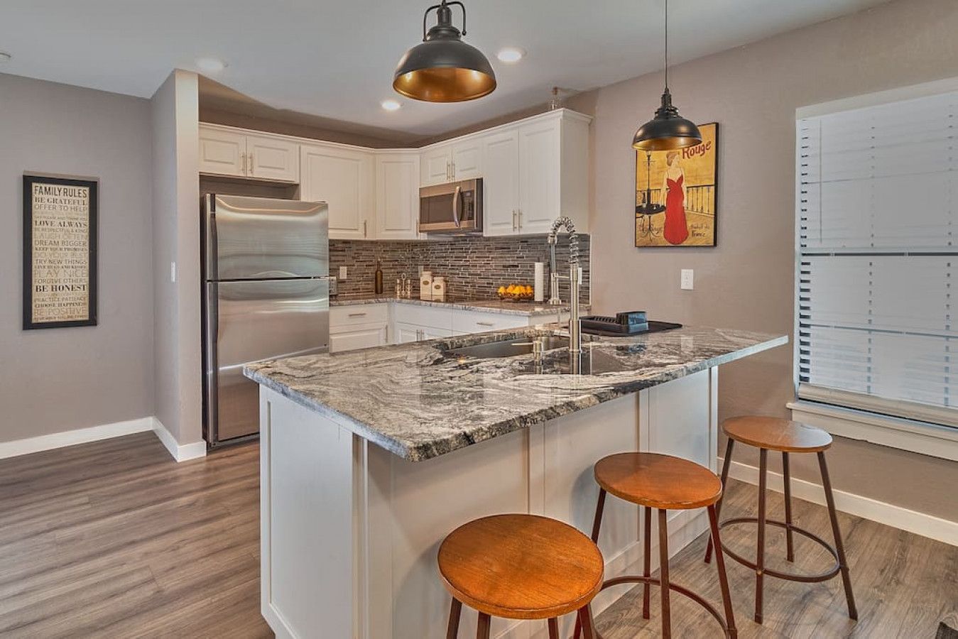A modern kitchen with white cabinets, a stainless steel refrigerator, a marble island with stools, and pendant lighting.