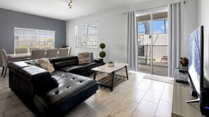 A modern living room with a sofa, coffee table, and TV. There are two windows with blinds, a balcony with a view, and a ceiling light fixture.