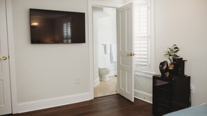 Une pièce avec un plancher en bois, un téléviseur noir fixé au mur, une porte blanche et un petit bureau noir sur lequel se trouve une plante.