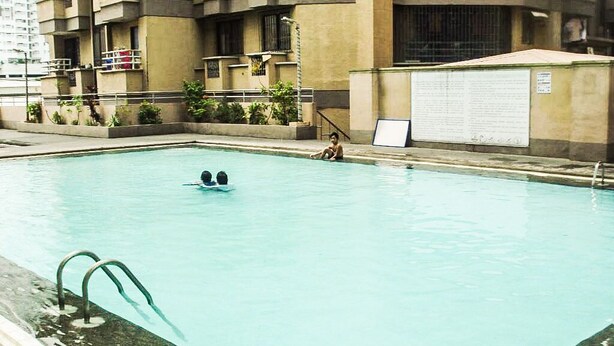 Una piscina con due persone che nuotano, circondata da un edificio con facciata in cemento e un cartello.