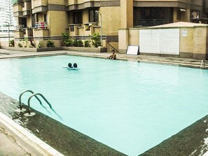 Une piscine où se trouvent deux personnes, entourée d’un bâtiment à l’extérieur en béton et sur lequel est apposée une enseigne.