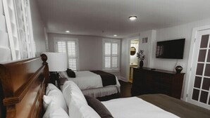 A hotel room with two beds, a wooden headboard, a TV, and a bathroom visible through an open door. - Southern Pines • The Jefferson Inn • Deluxe 2-Queen Suite (Southern Pines)