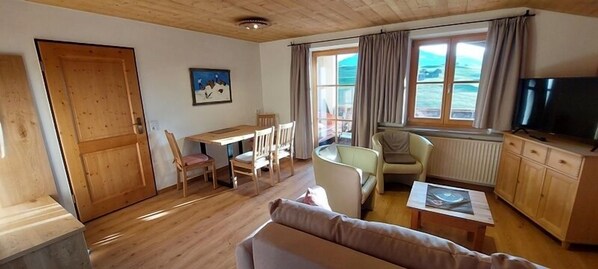 A cozy living room with a wooden floor, a sofa, a dining table with chairs, a TV, and a window with curtains. - Kaiserblick 2: Large two-room apartment with a spacious balcony (Reit im Winkl)