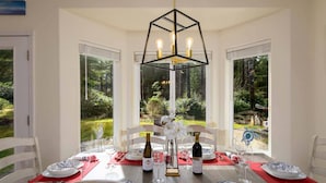 A dining area with a table set for six, featuring a geometric chandelier, white chairs, and a view of a garden through large windows. - New Listing! Beautiful well appointed Woahink lake area home w/ gas grill and fire pit (Florence)