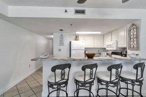 Una cocina con armarios blancos, un refrigerador, un microondas, un lavamanos y una barra con tres sillas.