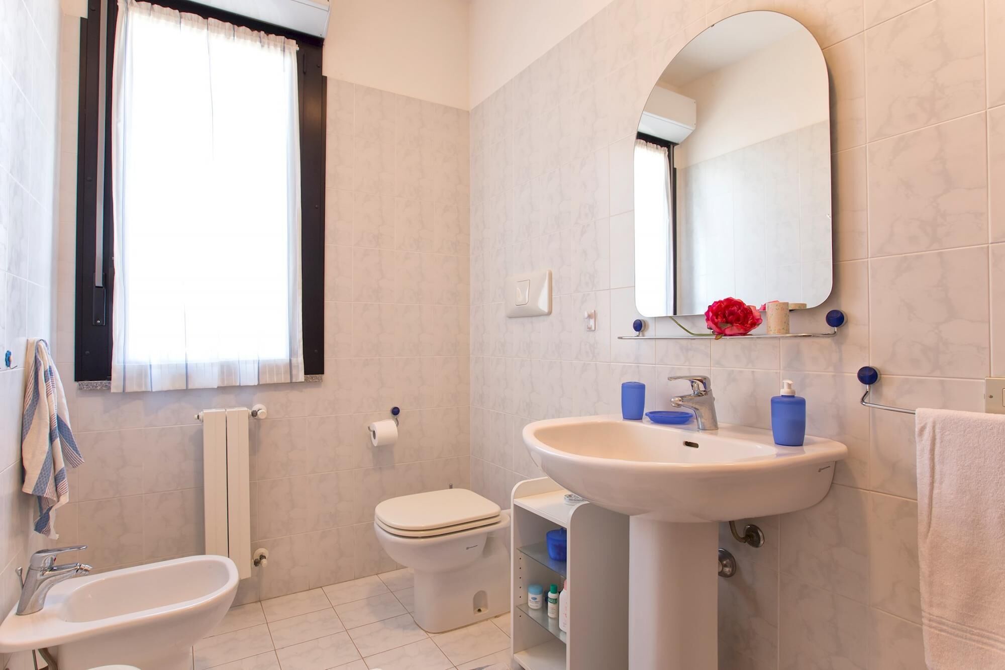 A bathroom with a toilet, sink, and bathtub. There is a window with curtains, a mirror, and a shelf with toiletries.