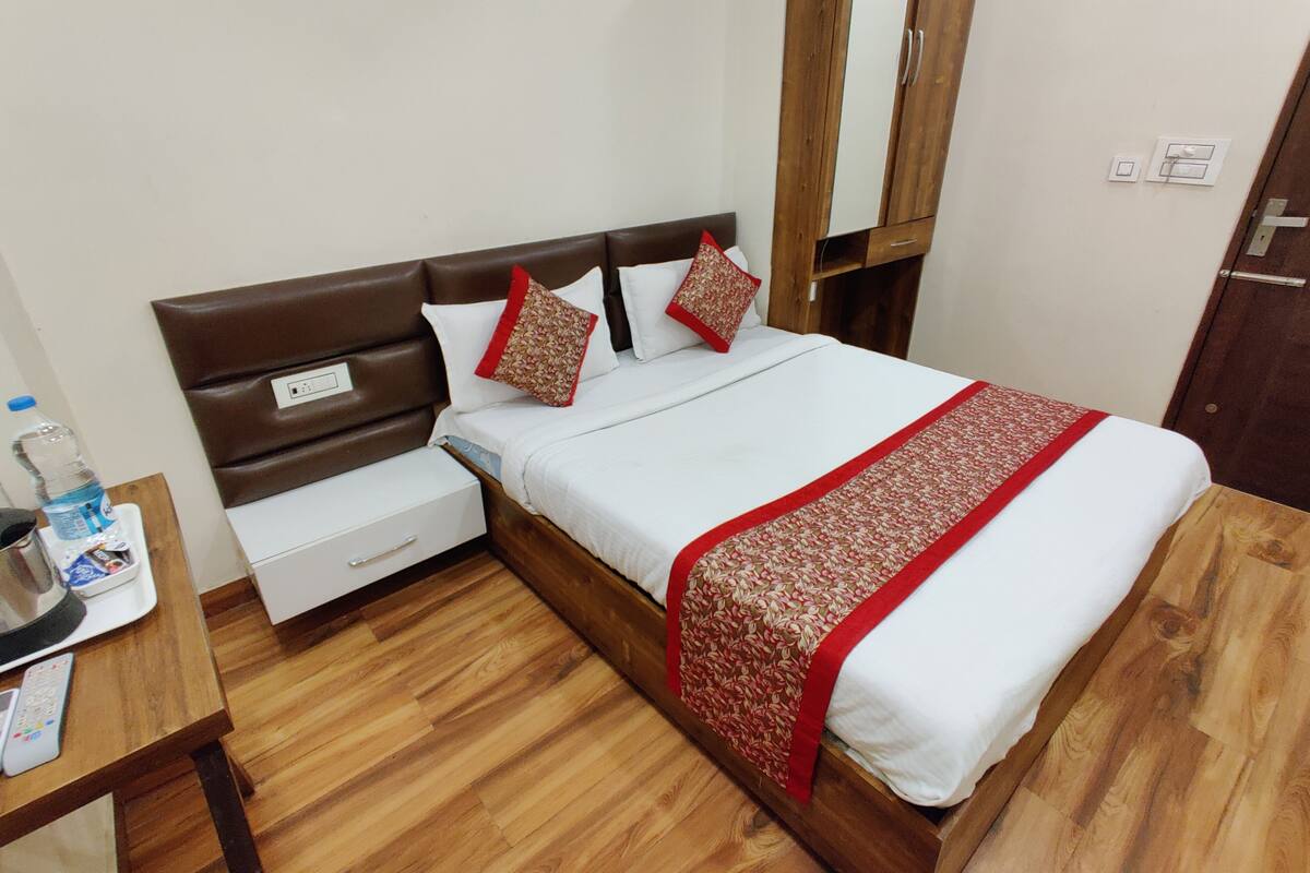 Una habitación de hotel ordenada con cabecera de madera, cama con ropa blanca y detalles rojos, mesita de noche con botella de agua y suelo de madera.